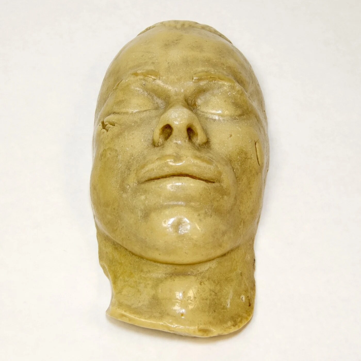 Pale yellow plaster death mask of a man's face, eyes closed, photographed from above on a white background.