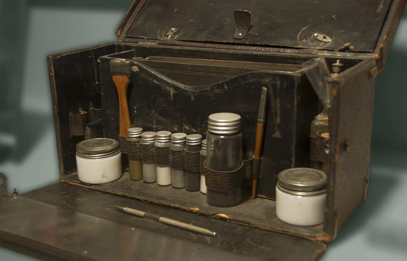 An open black case with a fitted tray holding jars, vials and a probe, with a hinged metal plate folded down below.