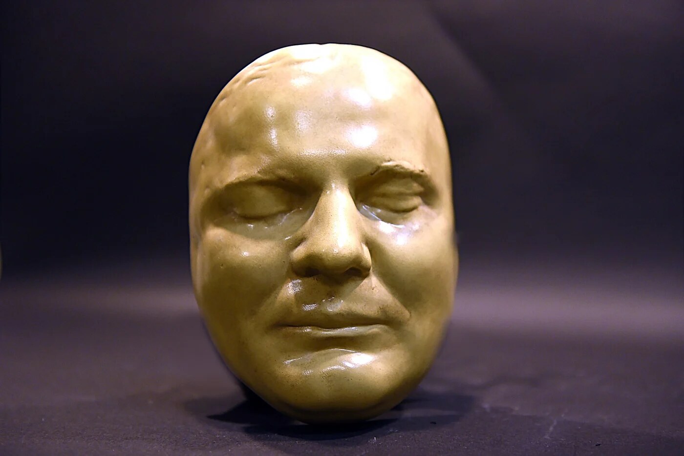Pale yellow mask of a man's face with closed eyes, photographed upright against a dark background.