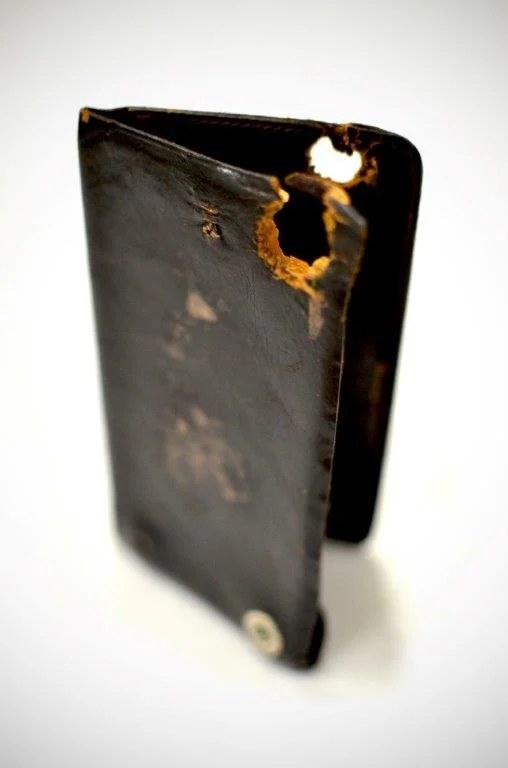 A dark leather notebook standing partly open against a white ground, with a ragged hole through the upper corner of its cover.