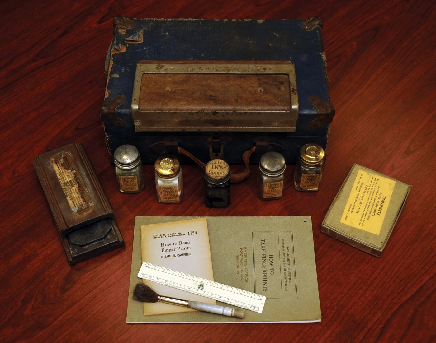 A paper booklet titled How to Take Fingerprints, from the Department of Justice, United States Bureau of Investigation, lying on a wooden surface with a second small booklet titled How to Read Finger Prints, an ink pad, a brush, a ruler, small bottles, a boxed packet and a closed case.