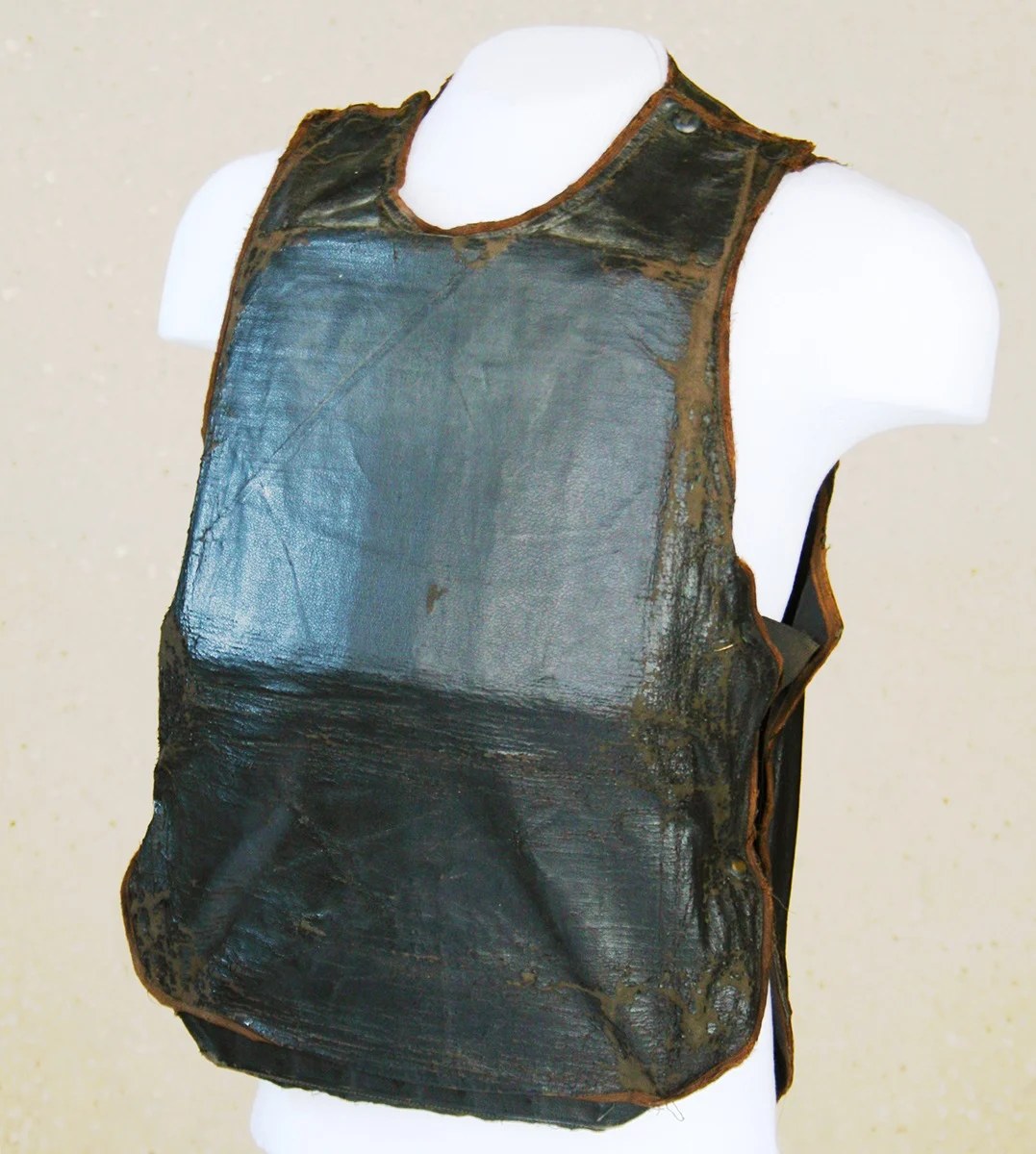 A dark, worn sleeveless vest with a cracked, shiny covering and brown edging, shown on a white mannequin torso.