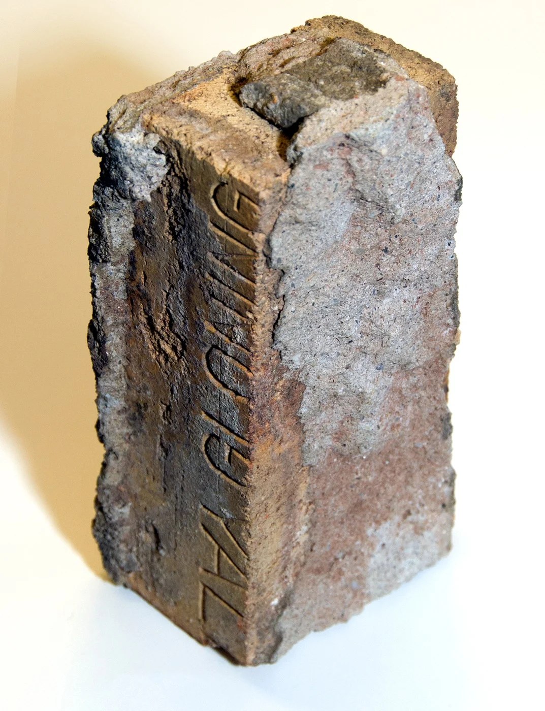A broken brick standing on end, with letters pressed into one face, photographed against a plain ground.