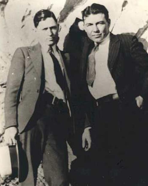 Sepia snapshot of two young men in suits and ties standing side by side in front of a rock face; the man at left holds a hat.