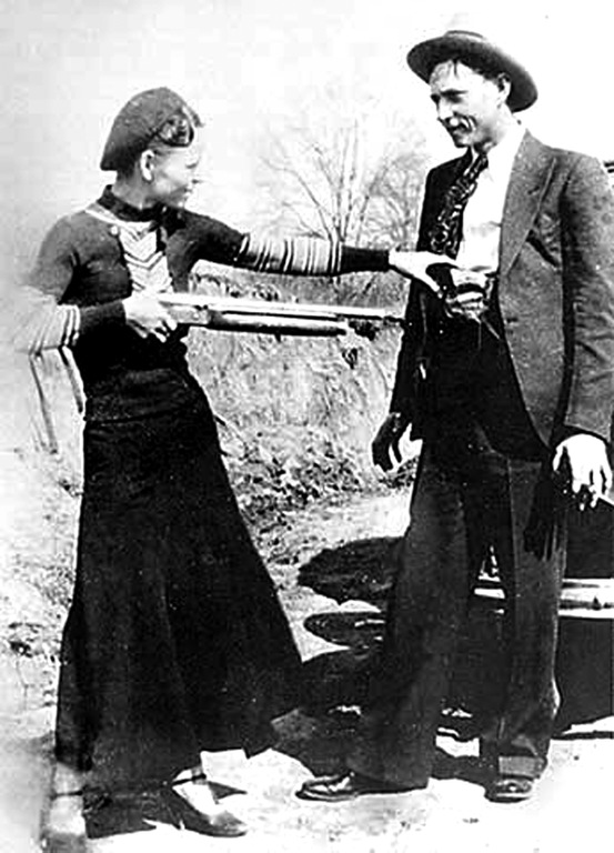 Black-and-white outdoor snapshot of a woman in a beret and long skirt holding a shotgun level toward a man in a hat and suit who holds a pistol at his side.