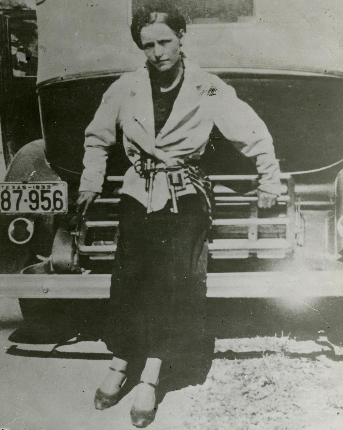 Black-and-white snapshot of Bonnie Parker in a light jacket and dark skirt, standing against the grille of a car whose license plate reads Texas 1933.