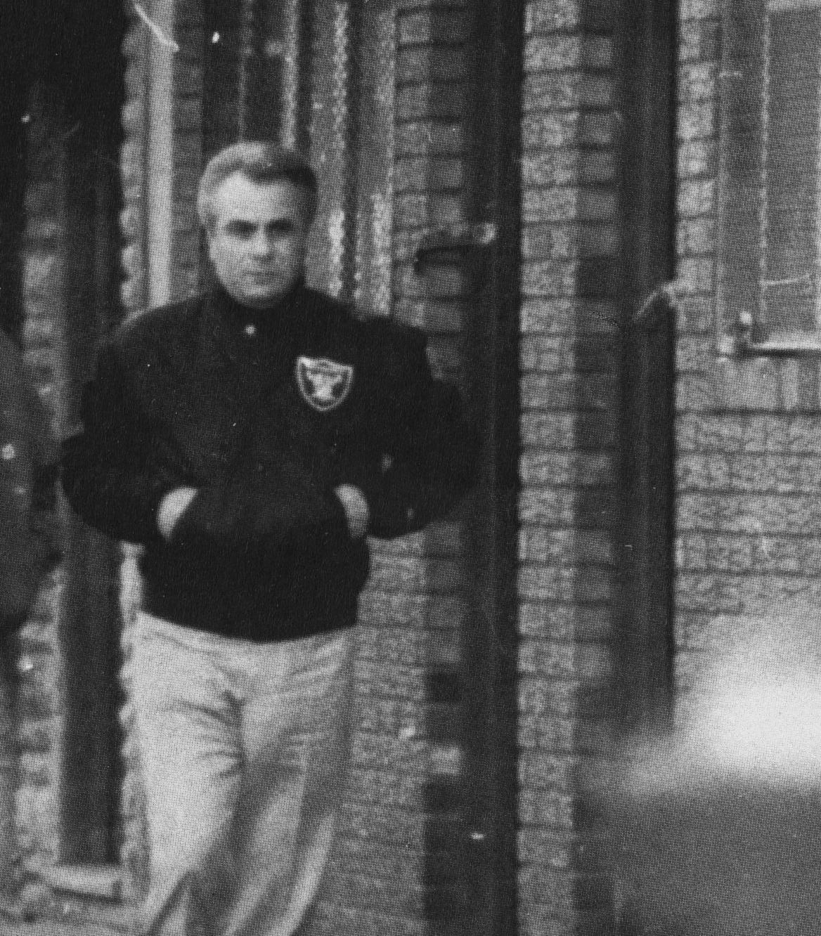 Surveillance photo of mobster John Gotti walking along a brick building, in a dark jacket with a team patch and light trousers, hands in his pockets.