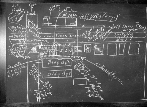 Chalk drawing on a blackboard of a street block with labels for Canal and Jeff Davis Parkway, boxes marked Bldg Apt, and handwritten notes for agents with shotguns and machine guns.