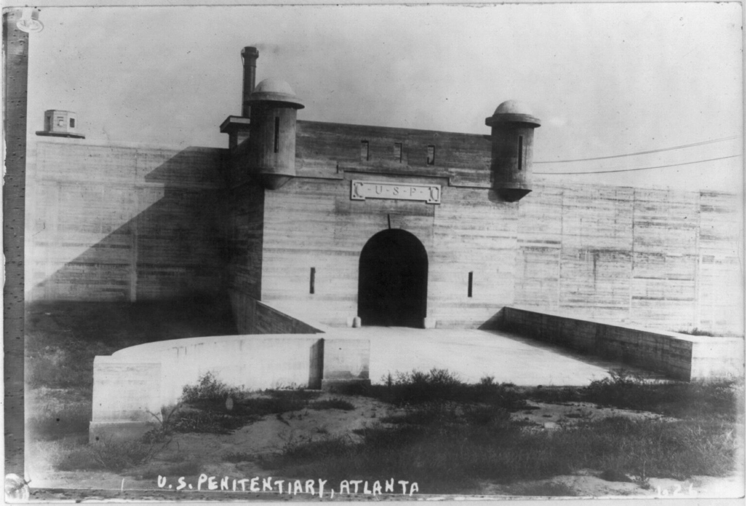 Black-and-white photograph of a walled gatehouse with two corner turrets and an arched entrance, with the words U.S. Penitentiary, Atlanta lettered at the lower edge.