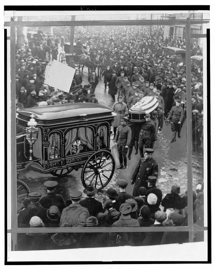 Funeral procession on a street lined with onlookers, with soldiers carrying a coffin behind a horse-drawn hearse.