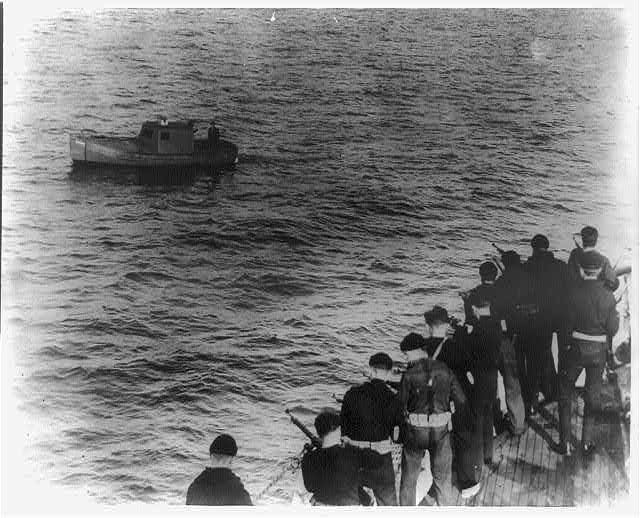 Armed men on the deck of a cutter aiming at a small boat on open water.