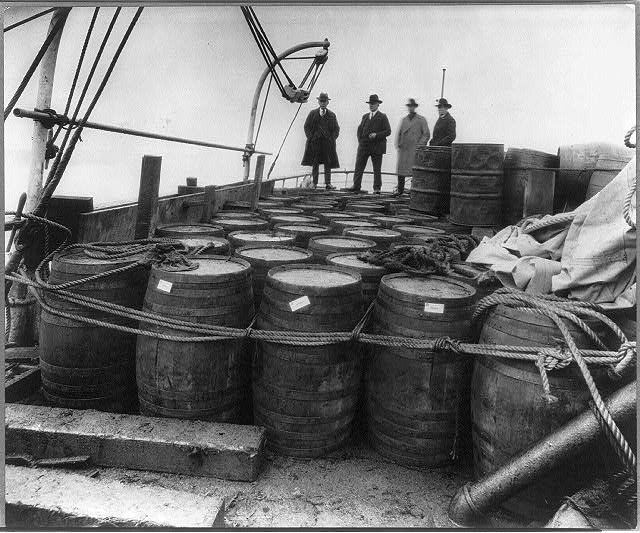 Men standing behind rows of barrels on the deck of a vessel.