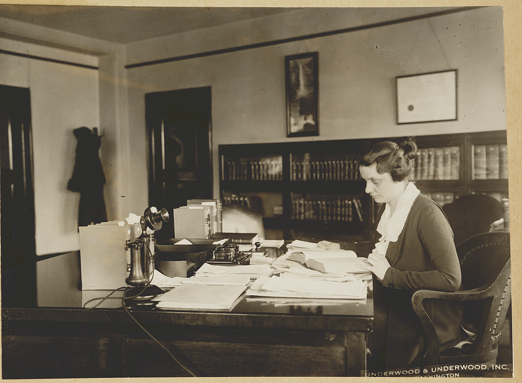 Mabel Willebrandt seated in her law office, reading a book at a desk covered with papers and books.