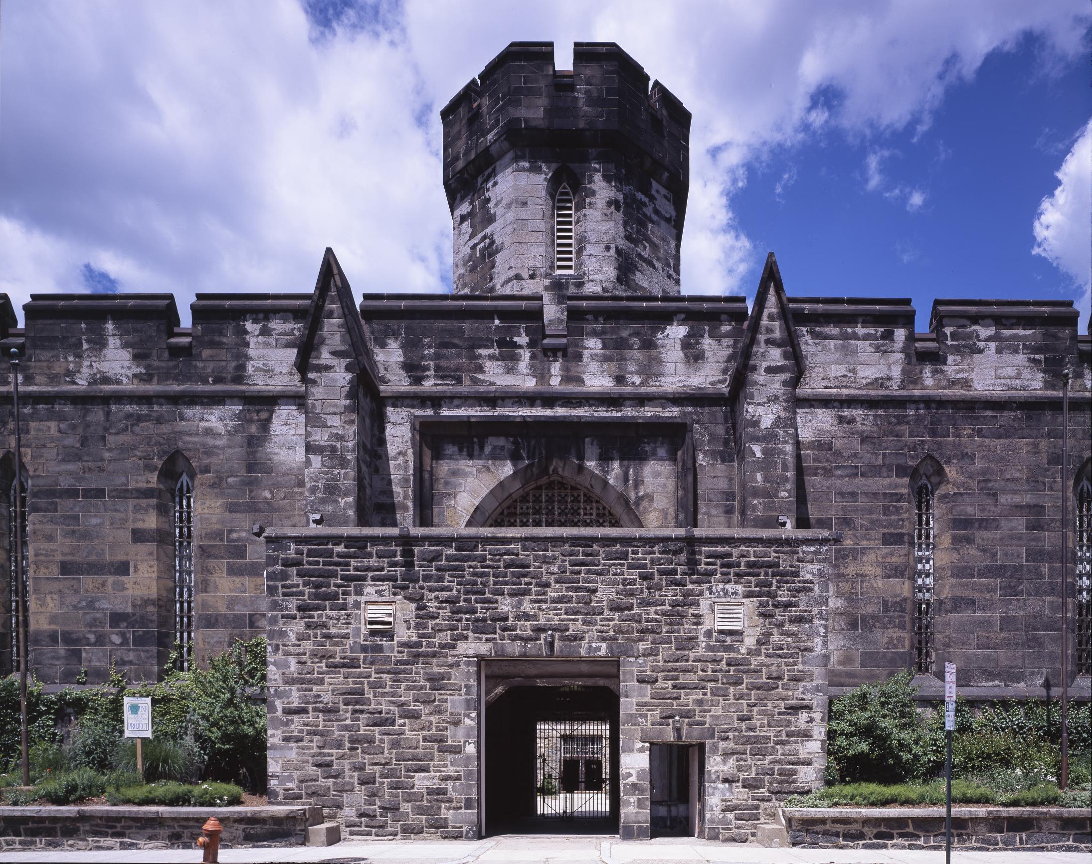 Color photograph of the gatehouse of Eastern State Penitentiary, an octagonal stone tower above a crenellated wall and a lower stone entrance block with an open gate and a barred inner gate.
