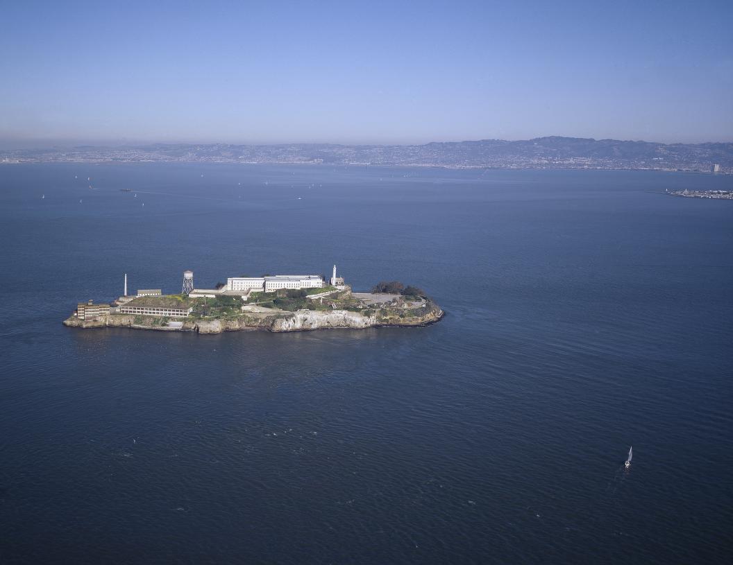 Color aerial photograph of Alcatraz Island in a blue bay, with buildings at the island's high point and a distant shoreline.