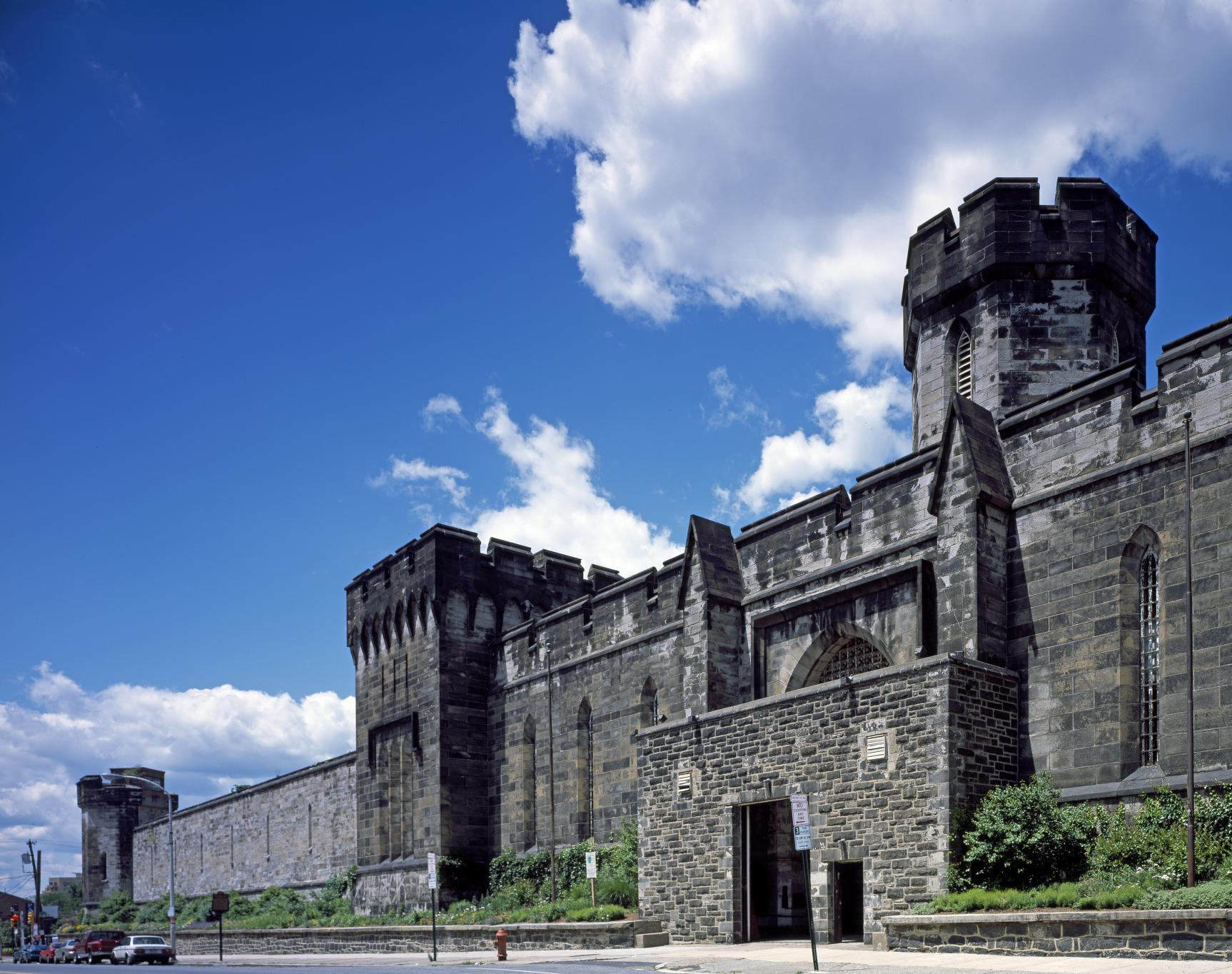 Color photograph of the stone front of Eastern State Penitentiary under a blue sky, with a crenellated tower above the entrance and a long wall running to a corner tower at left.