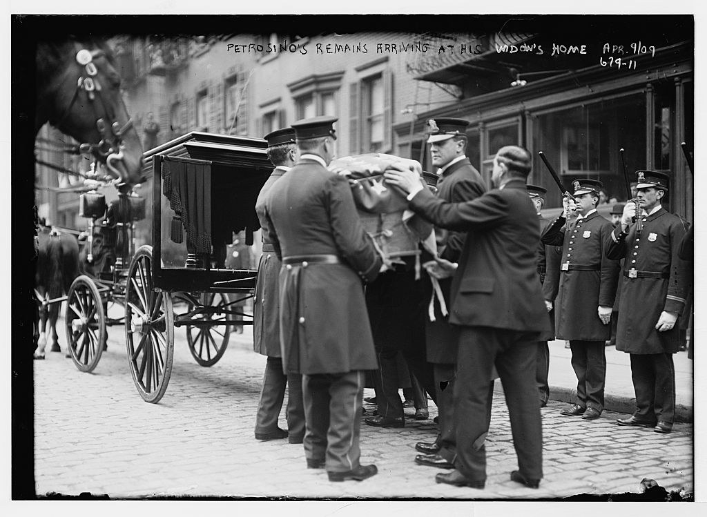 Police officers and men in overcoats lift a draped coffin from a horse-drawn hearse on a cobbled street while uniformed officers stand at attention; the negative is captioned 'Petrosino's remains arriving at his widow's home Apr. 9/09'.