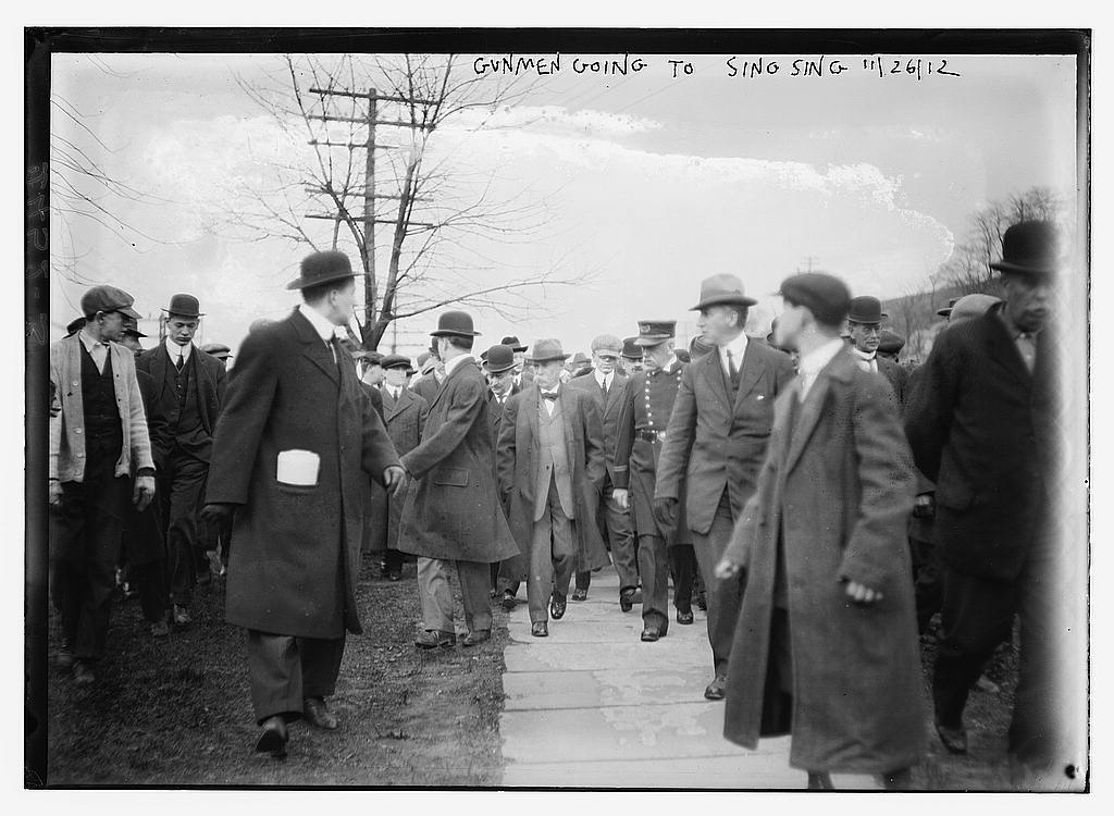Group of men in overcoats and hats walking along a path with escorts, November 1912.