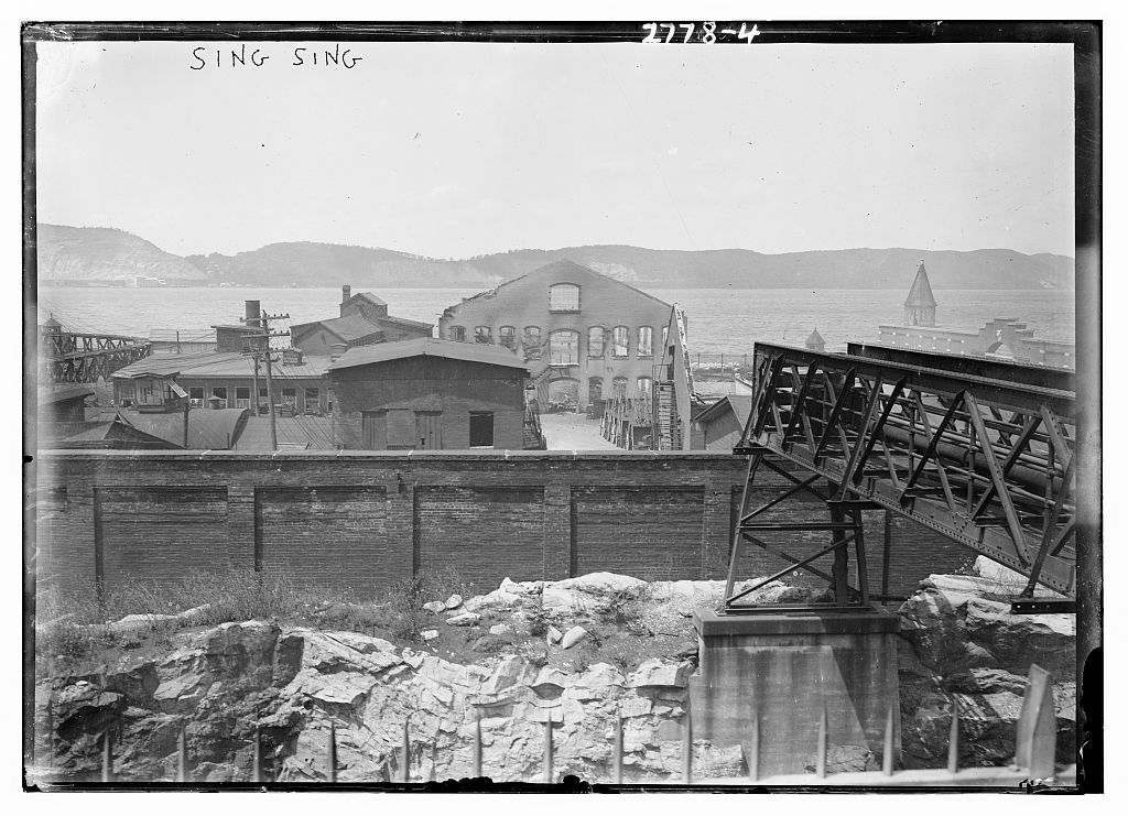 View of Sing Sing prison grounds with a wall, a bridge structure and the Hudson River and hills beyond.