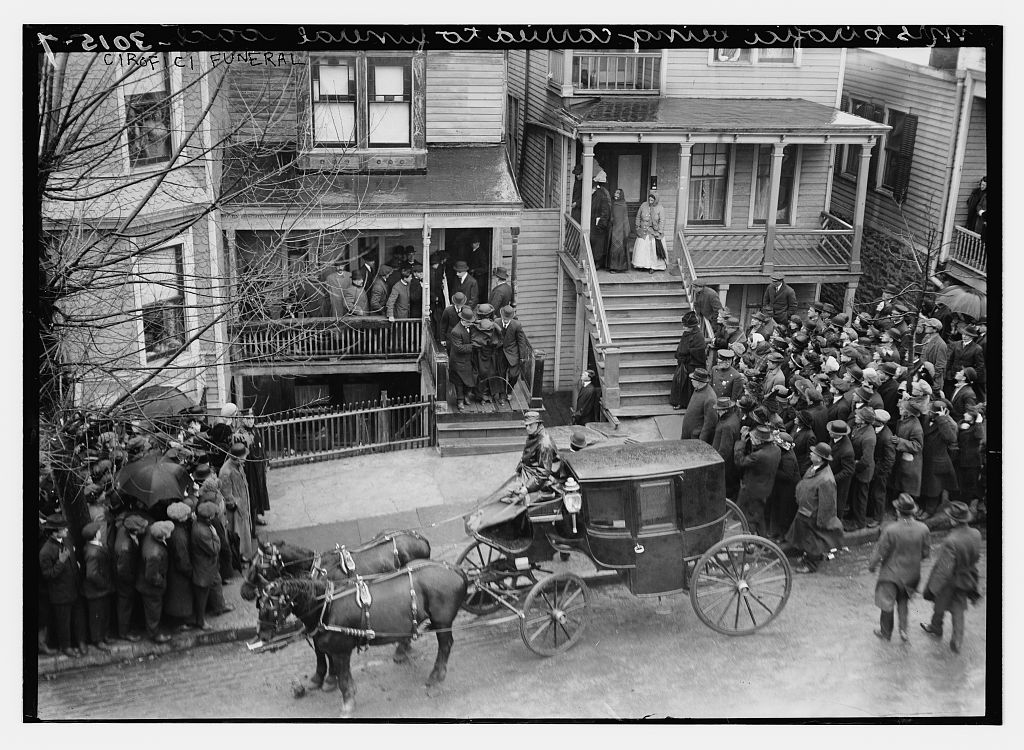 Crowd on a street in front of a wooden house as a horse-drawn funeral carriage waits, April 1914.