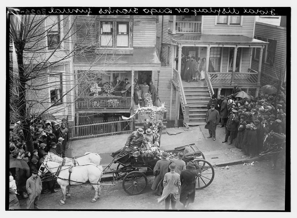 Horse-drawn funeral carriage carrying flowers outside a crowded house, April 1914.