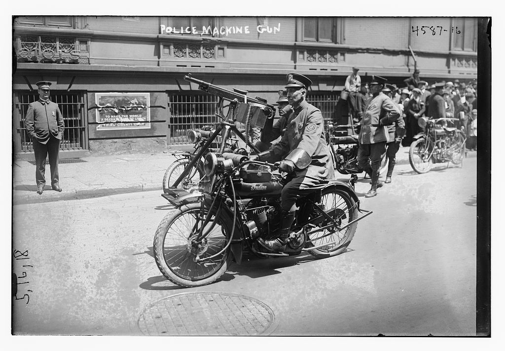 A police officer in uniform rides a motorcycle fitted with a mounted machine gun along a city street, with onlookers and other motorcycles behind; the words Police Machine Gun are lettered across the top of the negative.