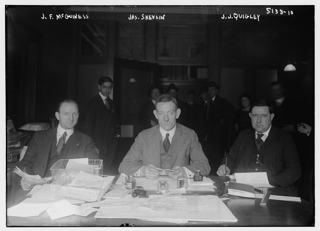 Three men in suits seated around a table covered with papers.