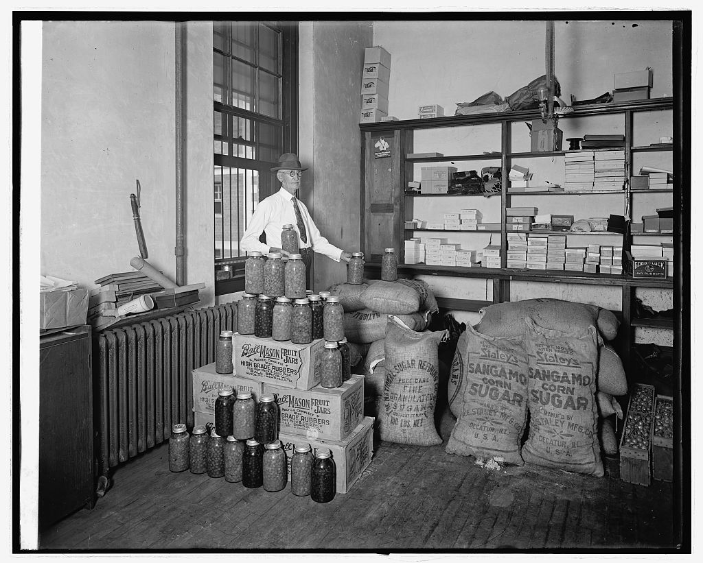 Man in a hat standing in a storeroom behind stacked fruit jars and sacks marked as corn sugar.