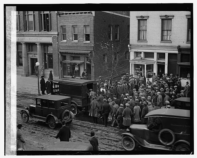 A crowd of men in hats surrounds a police patrol wagon on a street with streetcar tracks; parked automobiles stand in the foreground and a Quick Lunch sign hangs on the corner building