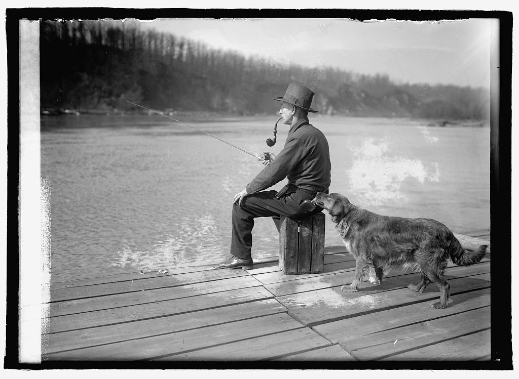 A dog sniffing a flask in the back pocket of a man seated on a pier with a fishing line over the Potomac River.