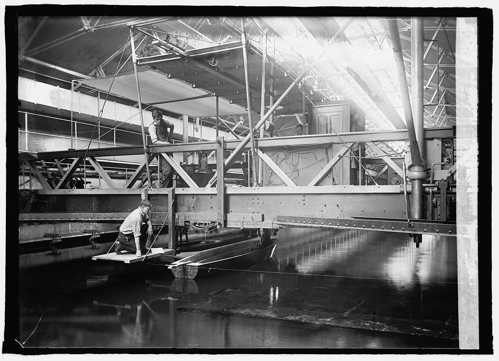 A man kneeling on a platform beside a small model boat in a test tank, with a second man on the overhead carriage.