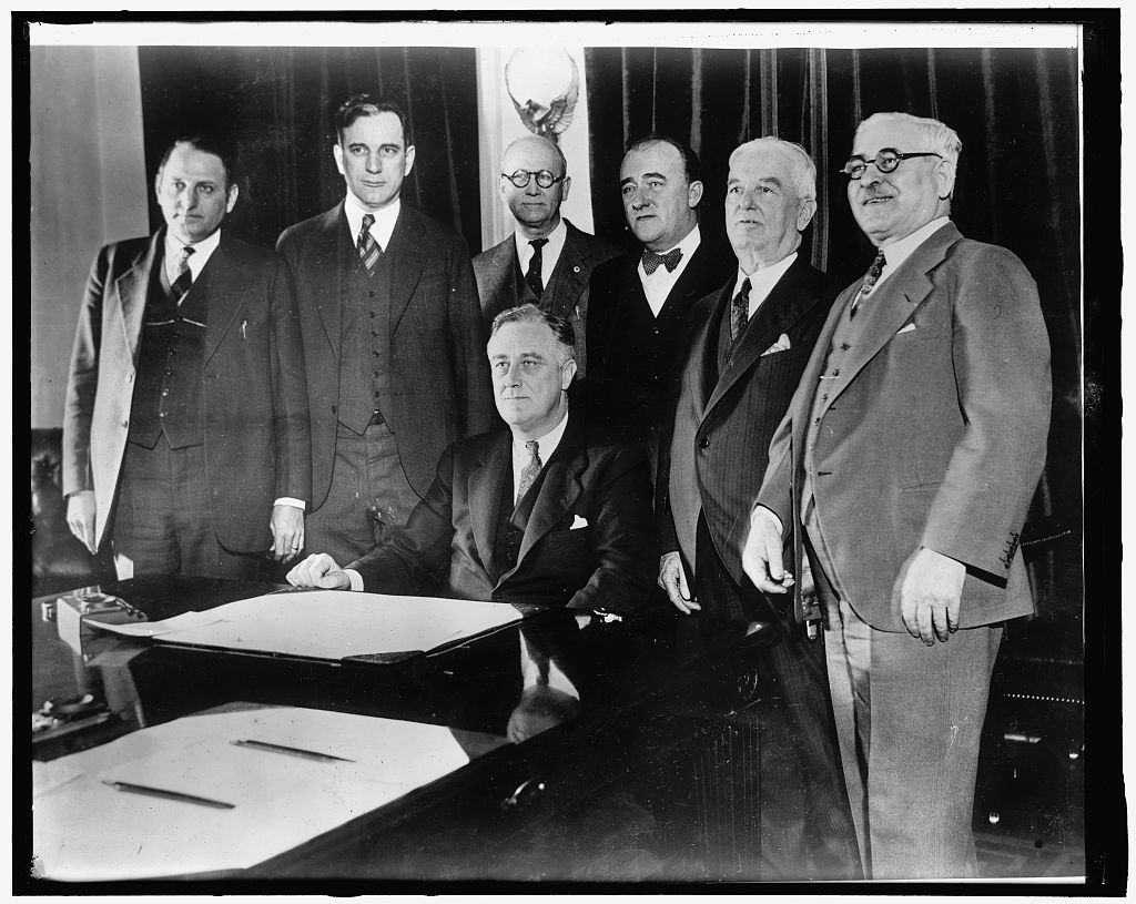 President Roosevelt seated at a desk signing a bill, with five men standing around him in the Cabinet Room.
