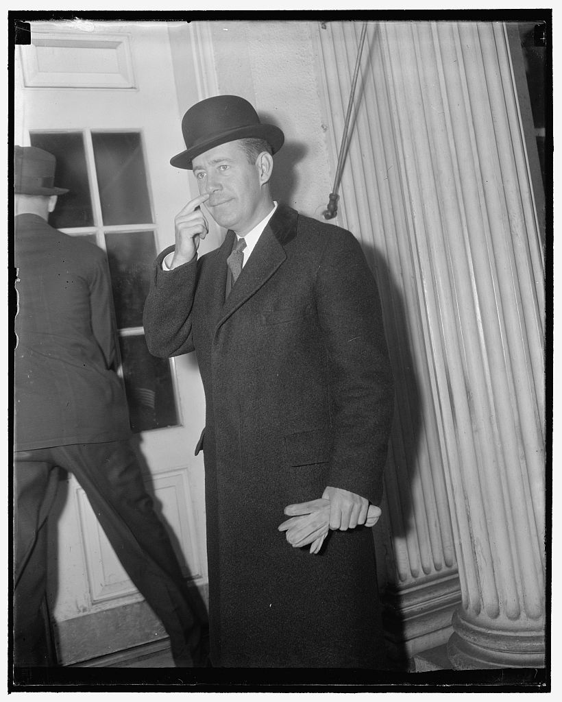 Herbert O'Conor in an overcoat and bowler hat, holding gloves, standing beside a white column at a White House entrance.