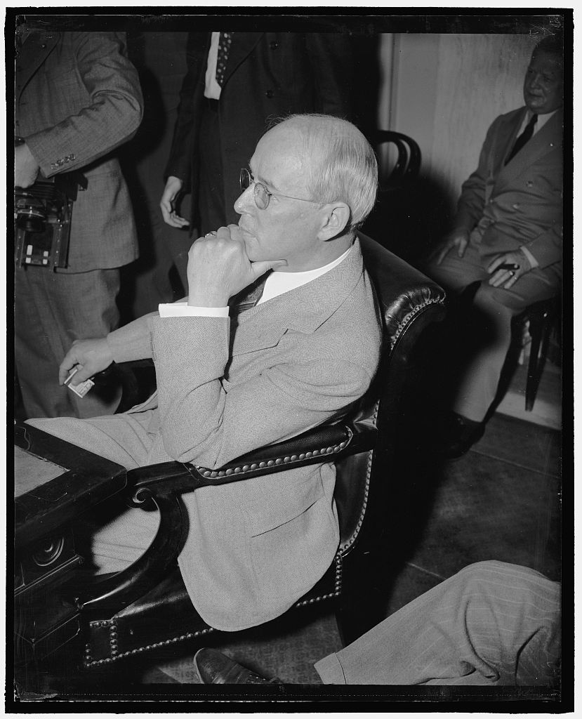 Senator Charles W. Tobey seated in a leather chair, chin on his fist, with other men standing and seated behind him.