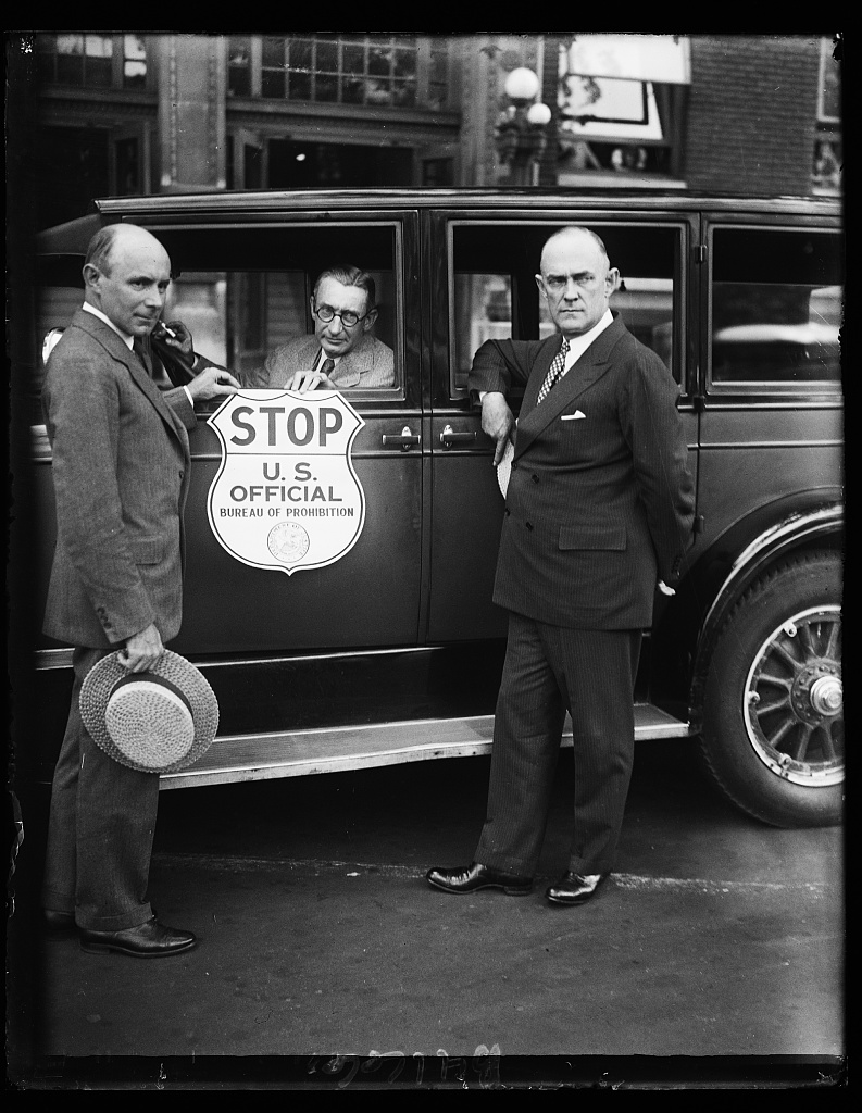Three men beside a car door bearing a shield-shaped sign reading STOP, U.S. Official, Bureau of Prohibition.