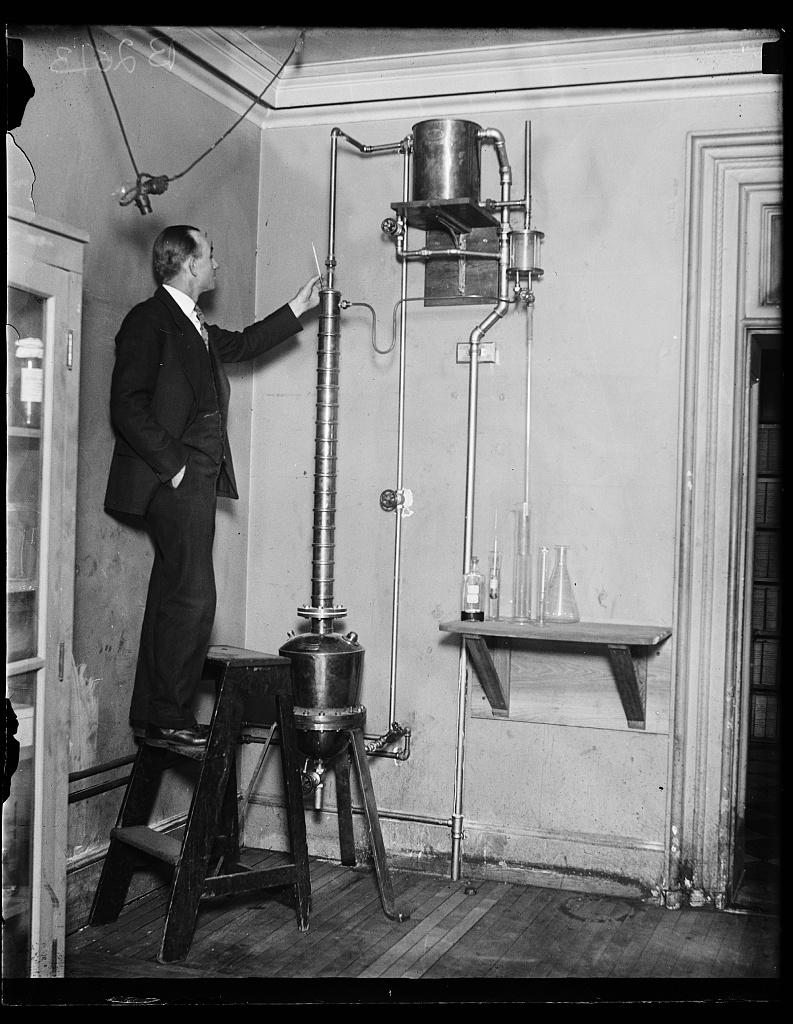 A man standing on a stool beside a small copper still in a laboratory.