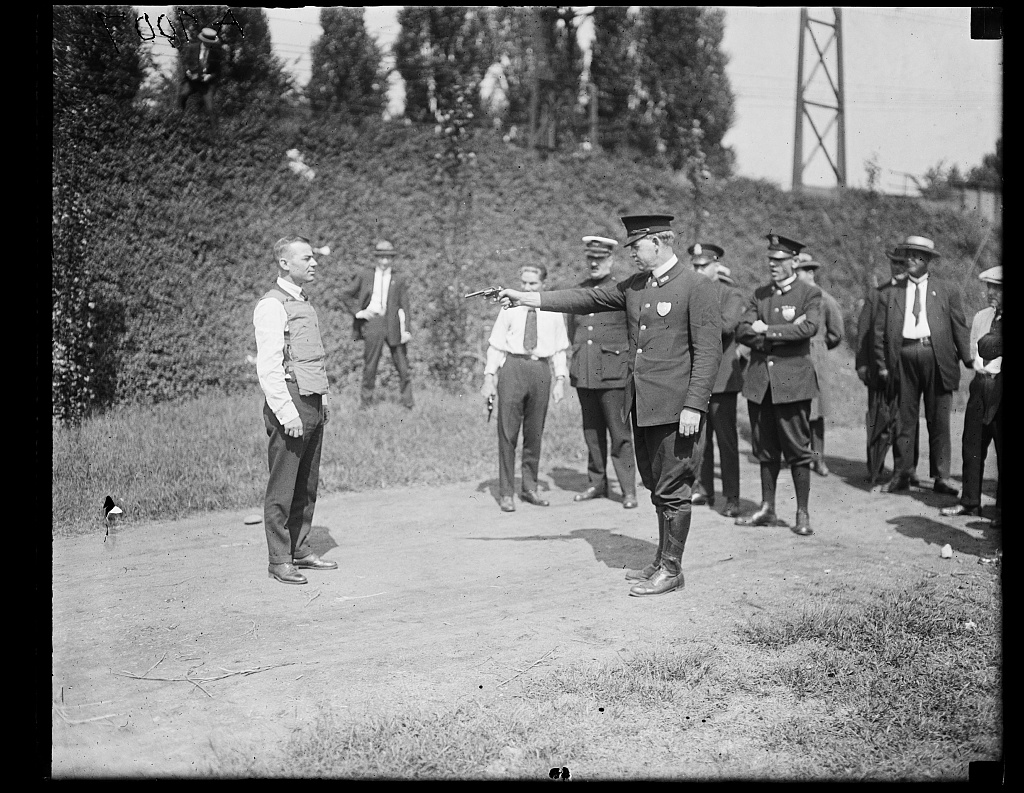 A man in a vest stands on open ground while a uniformed police officer points a pistol at him, with a group of officers and civilians watching.