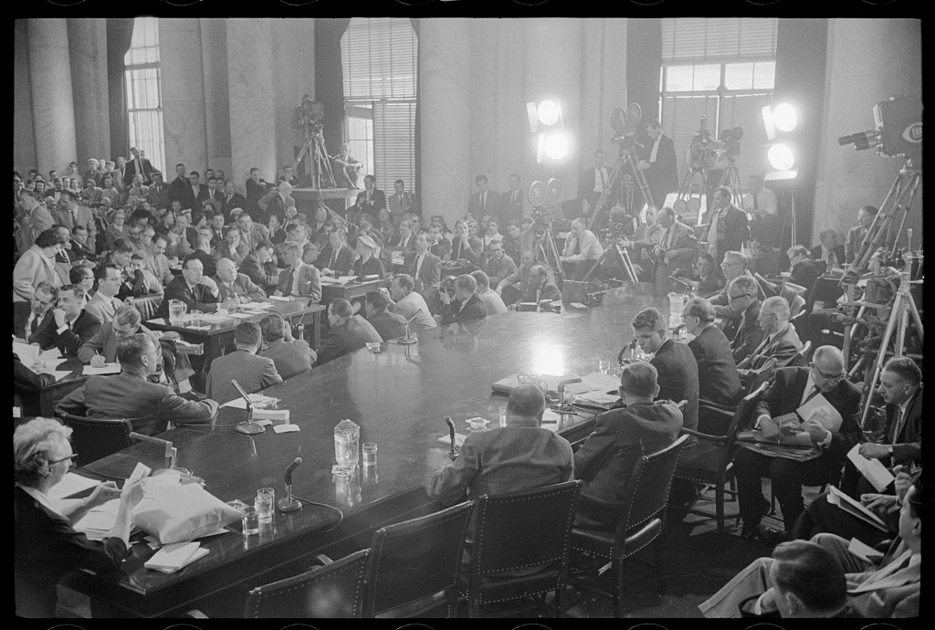 View across a committee table toward a packed hearing room with press lights and cameras at the back.