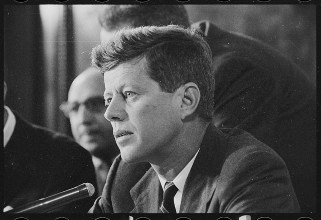 Senator John F. Kennedy in profile at a microphone during a committee session, with other men behind him, 26 February 1957.