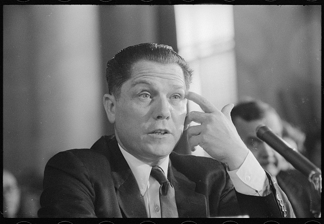 James R. Hoffa seated at a hearing table, touching his temple with two fingers, a microphone at right, 20 August 1957.
