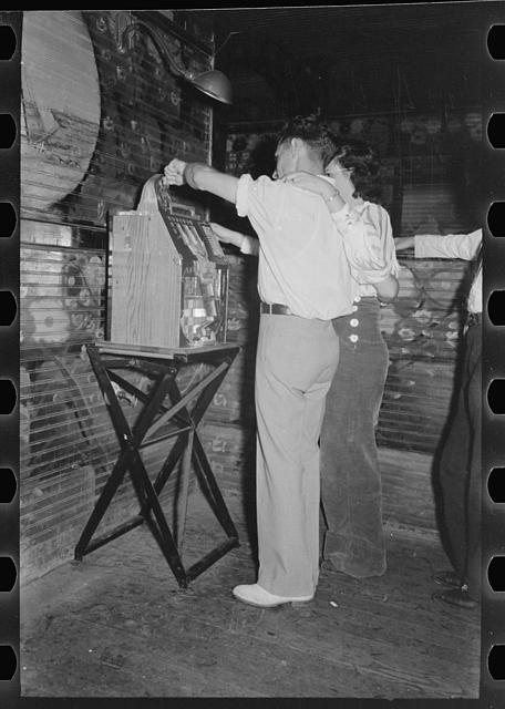 A man in a white shirt and light trousers pulls the handle of a slot machine on a folding wooden stand in a barroom with painted walls, a woman in a light blouse beside him and another person at the right edge