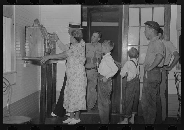 A woman in a flowered dress plays a slot machine mounted on a post in a white-walled room while a man in a straw hat, two other men and two barefoot boys watch