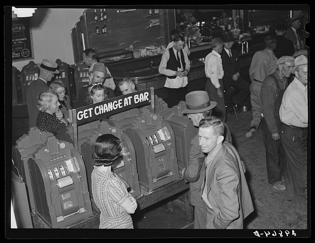 Men and women stand at a row of slot machines under a sign reading Get Change at Bar, with a long bar and more customers behind them