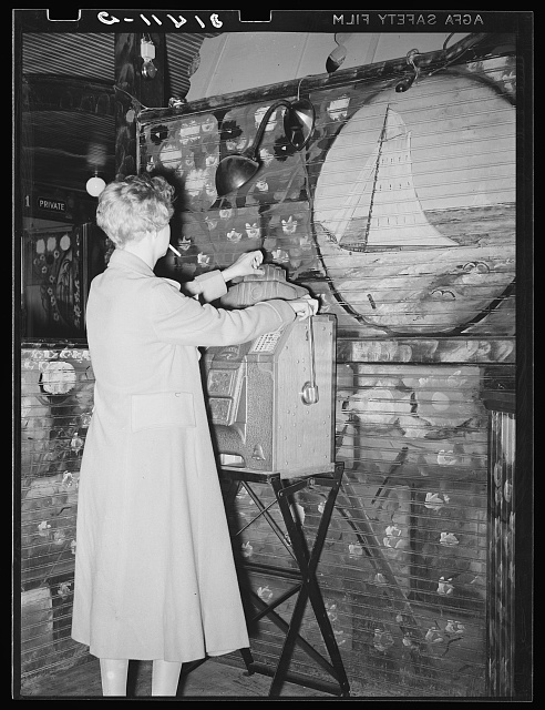 A person in a long light coat, a cigarette in the mouth, plays a slot machine on a folding stand in front of a wall painted with a sailing ship and flowers