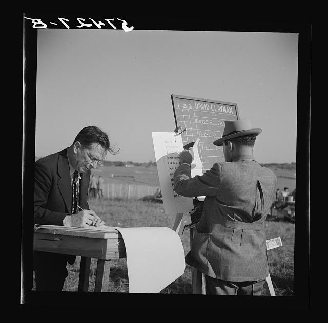 Outdoors on a grassy field, a man in glasses writes at a wooden table while a man in a hat and checked jacket stands at an easel holding a chalkboard of horse names