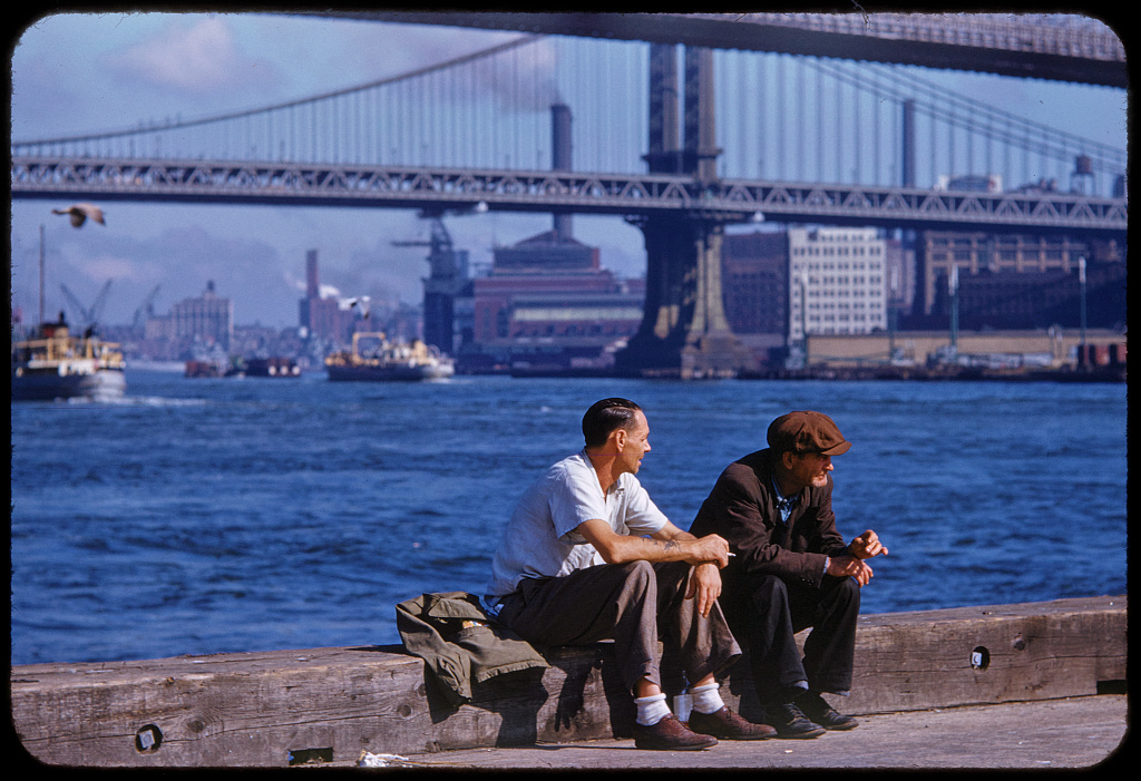 Two men seated on a wooden pier edge beside the East River with a bridge and boats behind them.