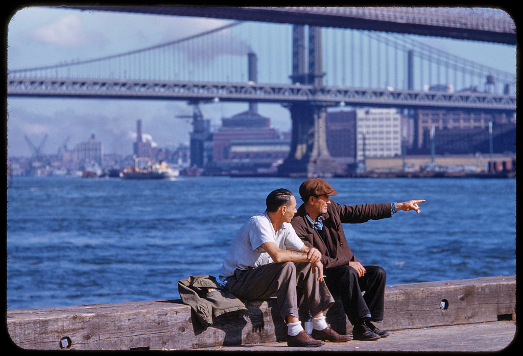 Two men seated on a pier beside the East River with a steel-towered suspension bridge in the background, one of them pointing across the water.