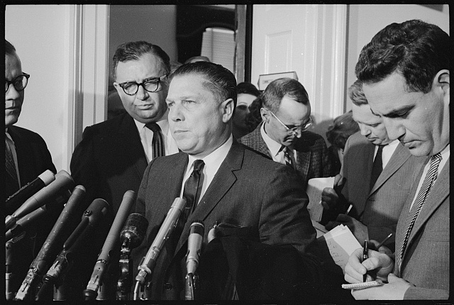 Jimmy Hoffa standing before a cluster of microphones in a doorway, with men taking notes around him, 1 February 1963.