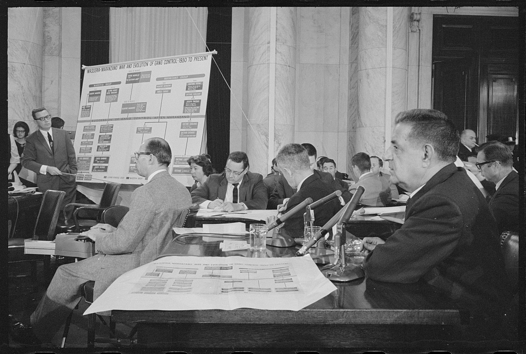 Joseph Valachi at a Senate subcommittee hearing while a man points to a chart, 1 October 1963.