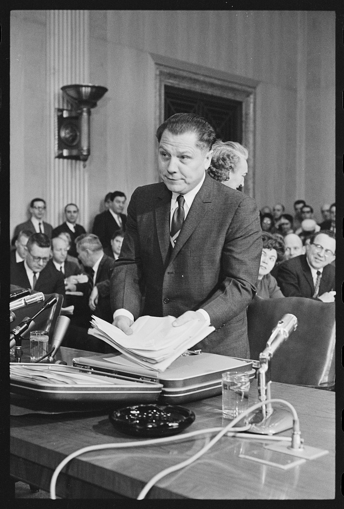 Man in a dark suit standing at a hearing table with a stack of papers in his hands, spectators behind him, January 1961.